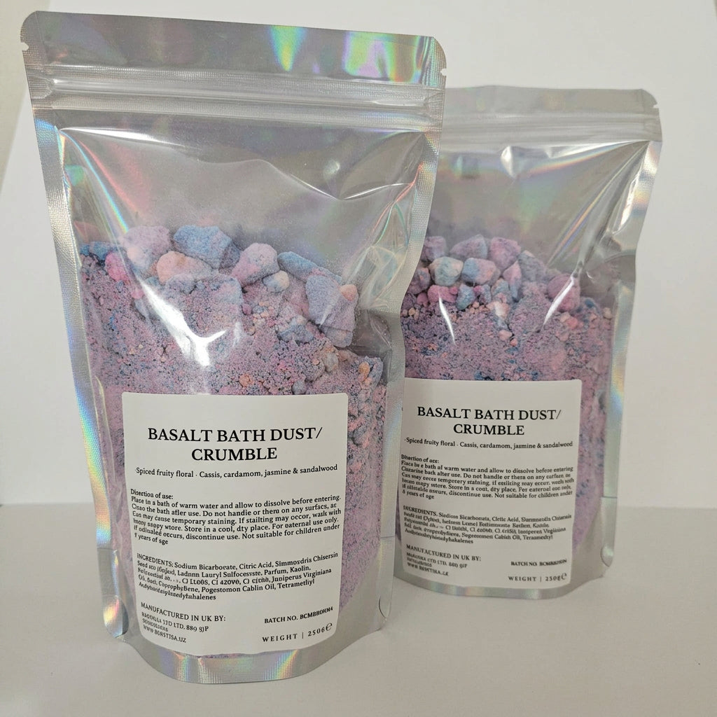 Two holographic stand-up pouches of Basalt Bath Dust and Crumble shown on a plain light background, with the front pouch fully visible and a second pouch partially behind it.
