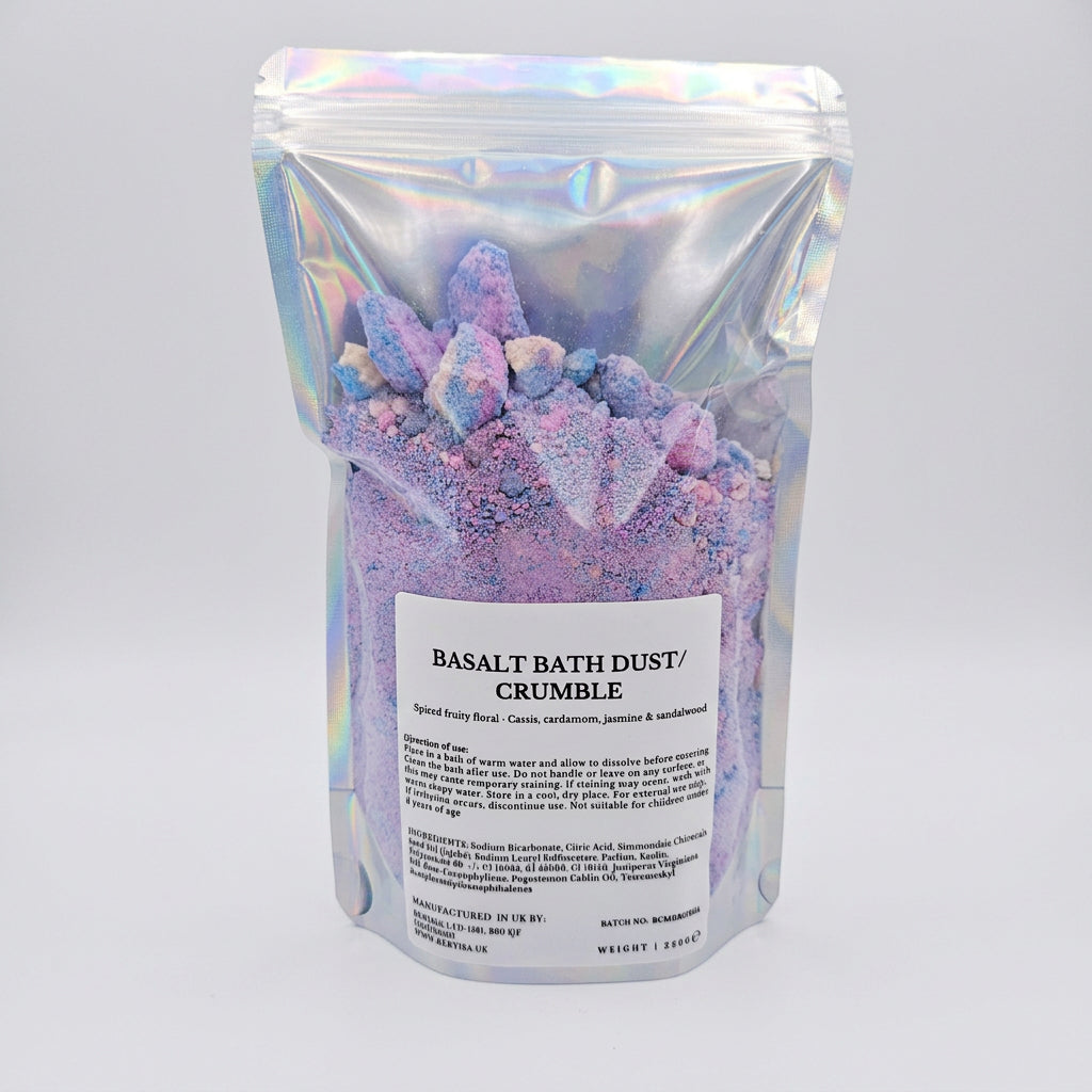 A stand-up pouch of Basalt Bath Dust / Crumble, filled with lavender and purple mineral powder and large crystal-like chunks, in an iridescent silver bag with a white label.