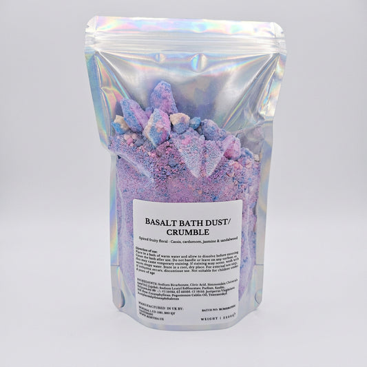 A stand-up pouch of Basalt Bath Dust / Crumble, filled with lavender and purple mineral powder and large crystal-like chunks, in an iridescent silver bag with a white label.