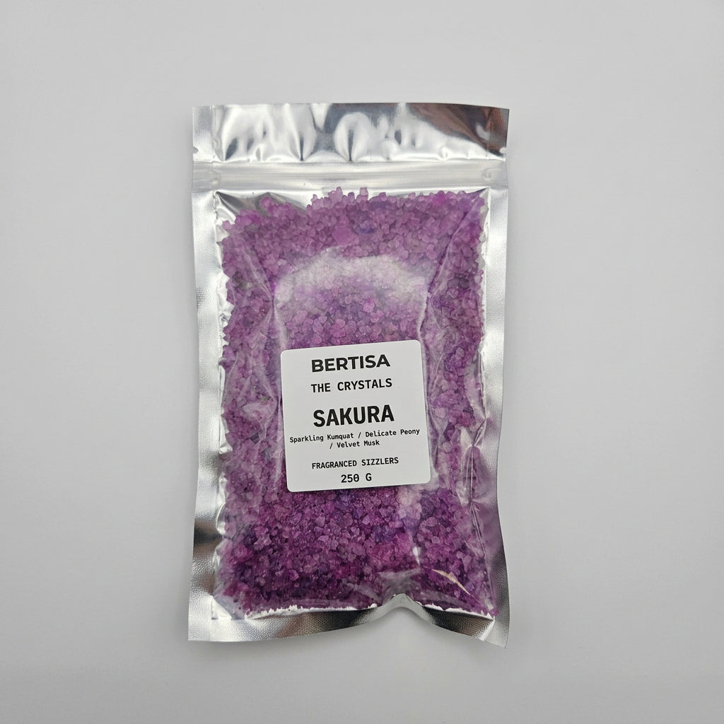 A silver resealable pouch containing bright purple crystalline salts, with a white label reading 'BERTISA THE CRYSTALS SAKURA FRAGRANCED SIZZLERS 250 G'.