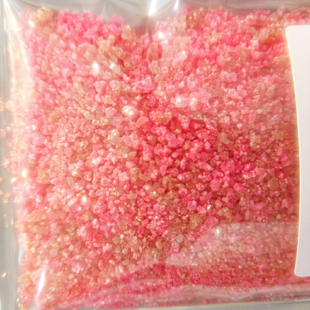 A close-up of bright pink and peachy-pink coarse crystals inside a clear plastic bag, with a white label partially visible on the right side.