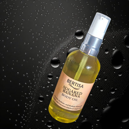A clear spray bottle labeled 'BERTISA SUGARED BANANA BODY OIL' with a creamy yellow liquid visible inside, presented against a dark background with water droplets around it.